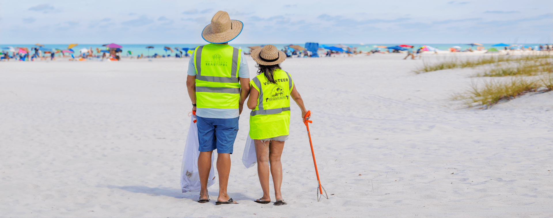 a man and woman in Keep Manatee Beautiful and Keep America Beautiful volunteer reflective vests, holding a trash picker on an anna maria island beach