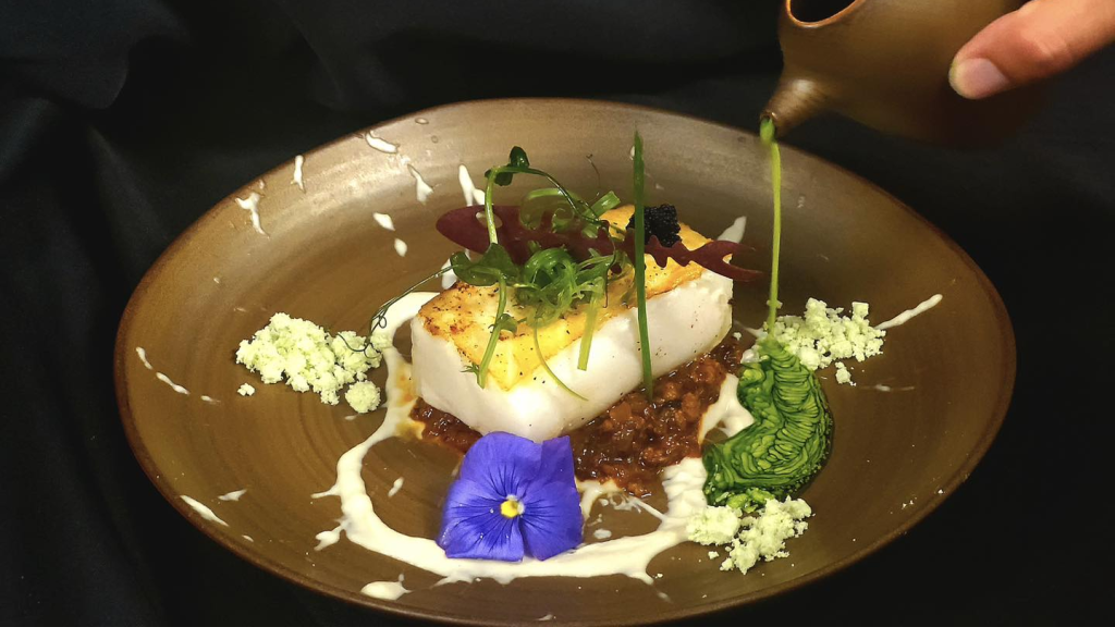 a fancy dish on a black background with several small food items an a hand pouring a sauce 