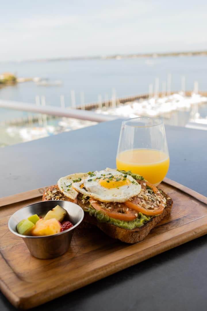 a sandwich with a egg and a bowl of fruit on a wooden board with a view of a marina